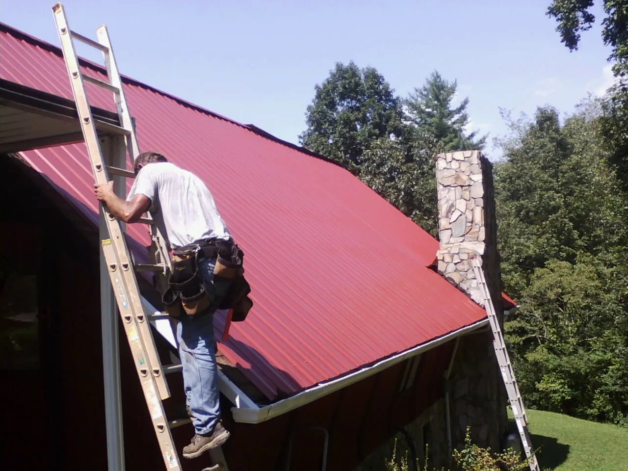 Roofer on a ladder beside a red metal roof and stone chimney — Soffit & Fascia Repair by Roofing Brothers in Hitchcock